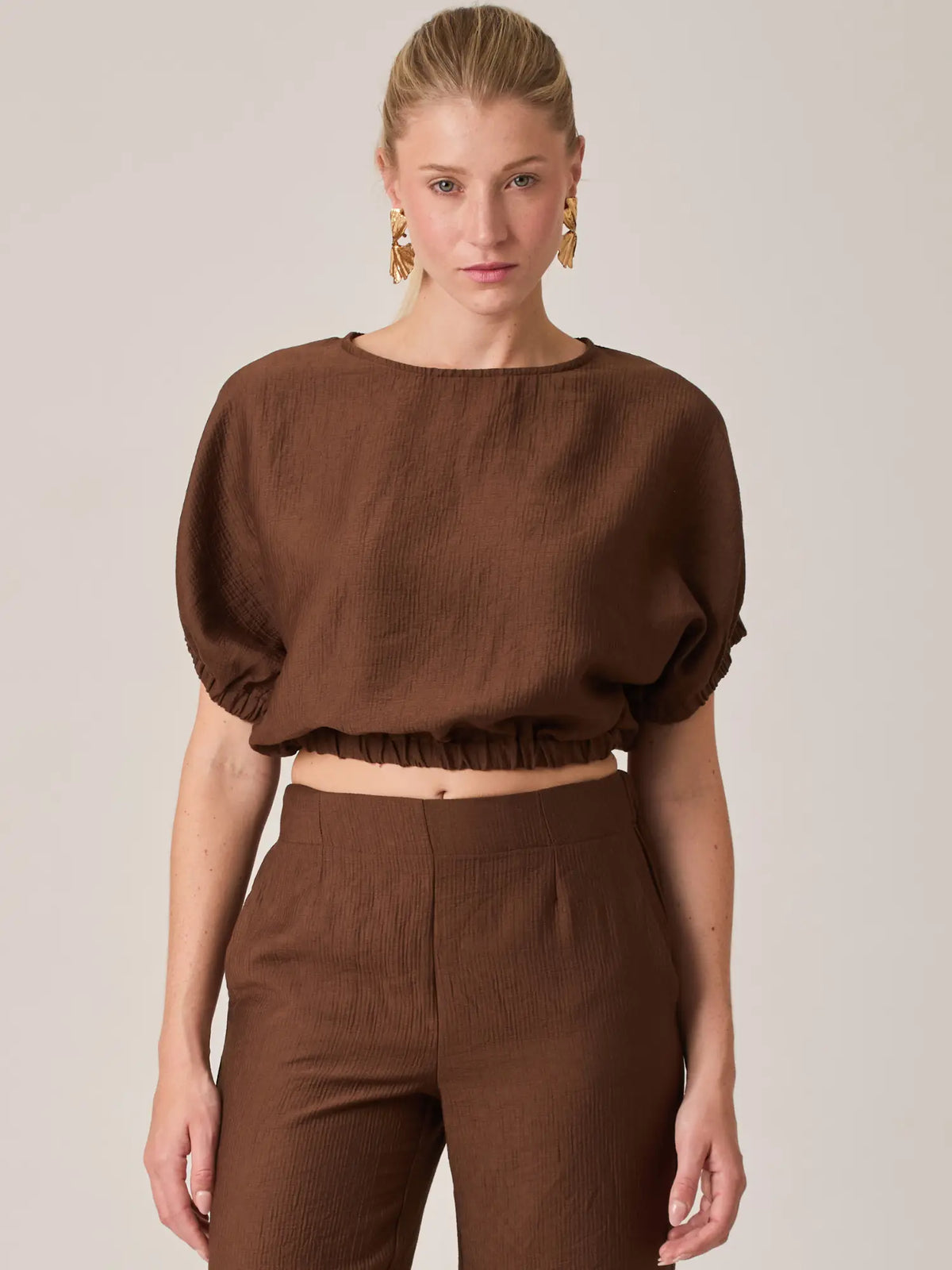 La Blusa Agostina Café es una prenda femenina de diseño moderno con manga corta y ajuste elástico en la cintura que realza la silueta. Ideal para looks casuales o elegantes, perfecta para combinar con pantalones de tiro alto.