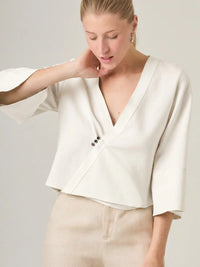 Blusa cruzada blanca con escote en V y boton decorativo que aporta un toque sofisticado. Ideal para outfits de dia o eventos casuales, esta blusa combina comodidad y elegancia.