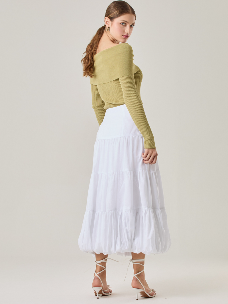 Esta es una falda blanca de largo midi con un diseño en capas que crea un efecto abombado en la parte inferior. Está hecha en una tela liviana y con caída. Tiene un estilo romántico y femenino, ideal para combinar con blusas ajustadas o crop tops. En la imagen se lleva con un suéter off-shoulder que le da un look sofisticado.