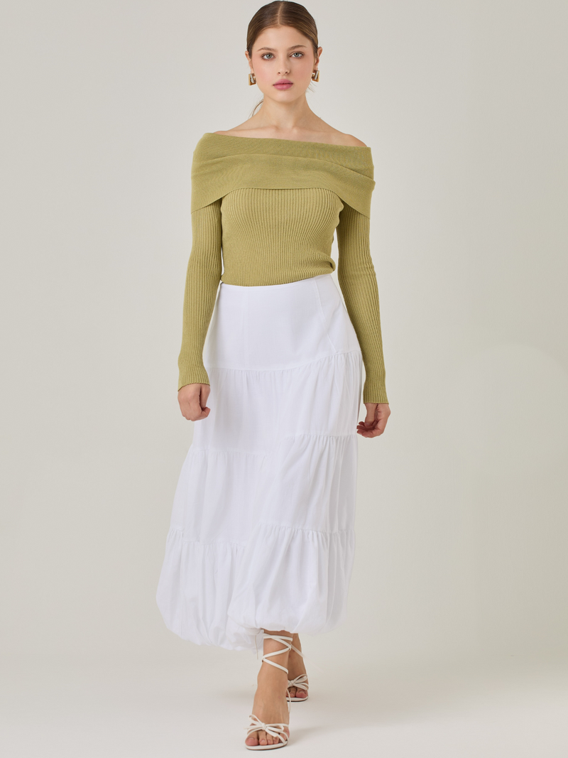 Esta es una falda blanca de largo midi con un diseño en capas que crea un efecto abombado en la parte inferior. Está hecha en una tela liviana y con caída. Tiene un estilo romántico y femenino, ideal para combinar con blusas ajustadas o crop tops. En la imagen se lleva con un suéter off-shoulder que le da un look sofisticado.