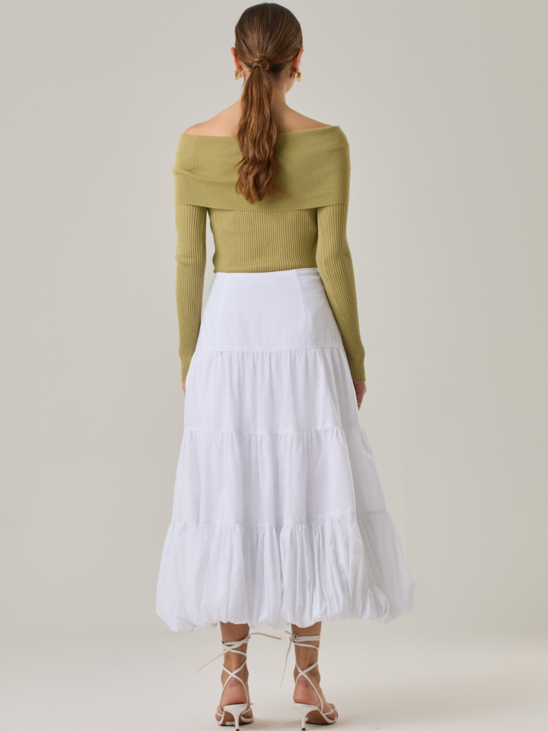 Esta es una falda blanca de largo midi con un diseño en capas que crea un efecto abombado en la parte inferior. Está hecha en una tela liviana y con caída. Tiene un estilo romántico y femenino, ideal para combinar con blusas ajustadas o crop tops. En la imagen se lleva con un suéter off-shoulder que le da un look sofisticado.