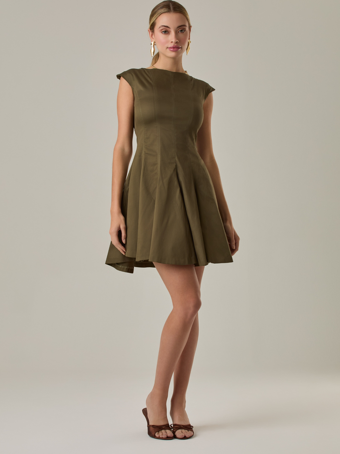 Vestido corto con silueta entallada en la parte superior y falda amplia. Tiene cuello cerrado tipo caja, mangas pequeñas que apenas cubren los hombros y costuras que definen la estructura del torso. La falda incorpora pliegues profundos que le dan volumen y movimiento. El largo llega por encima de la rodilla. Cierre invisible en la espalda.
