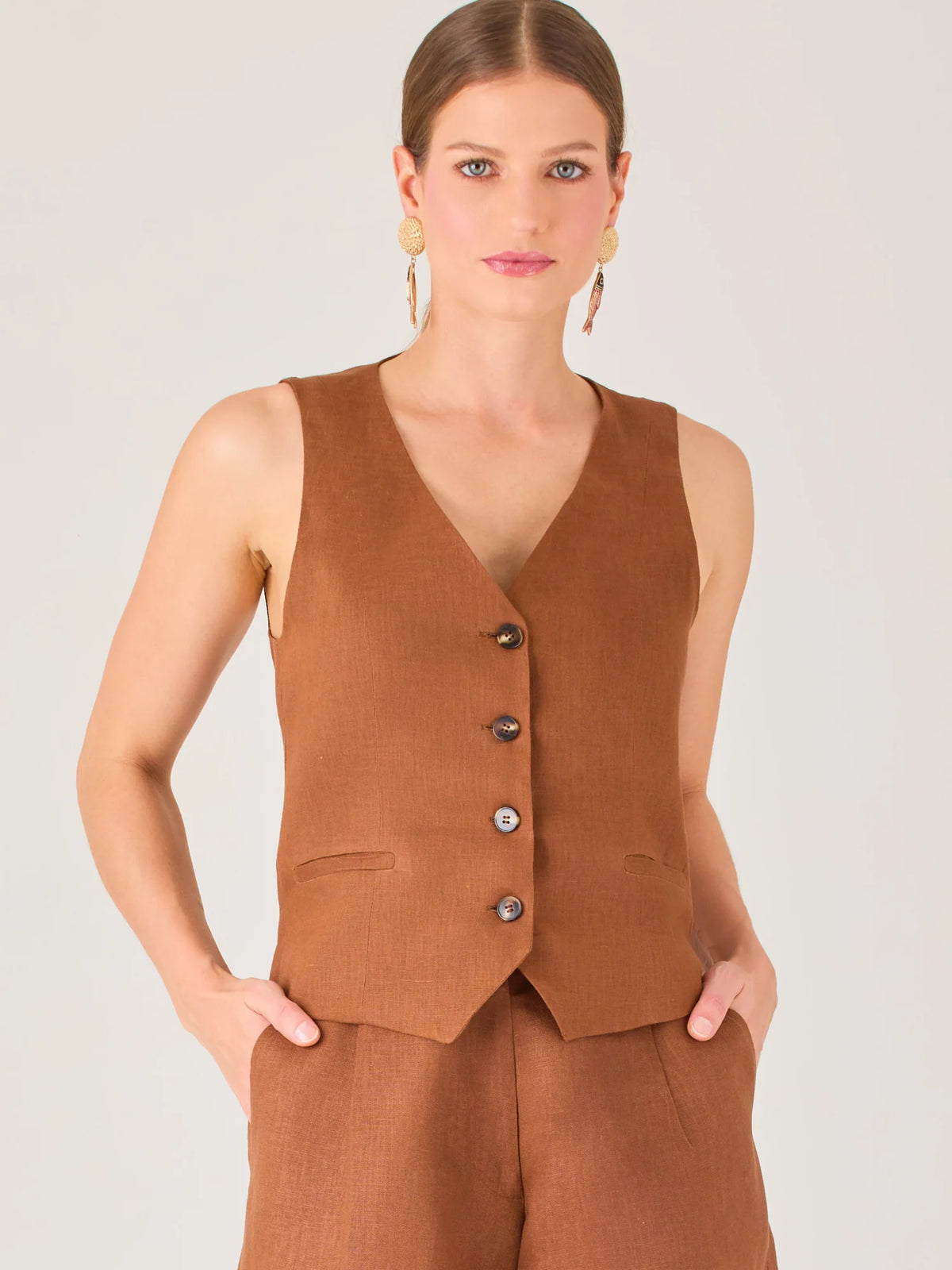 Esta blusa tipo chaleco es ideal para climas cálidos o para crear looks sofisticados con un aire casual. Presenta un corte recto ligeramente entallado que acentúa la silueta de forma sutil, y un escote en “V” que estiliza el cuello. Cuenta con una botonadura frontal compuesta por cinco botones oscuros que contrastan elegantemente con el tono neutro de la prenda. También incorpora bolsillos falsos decorativos a ambos lados del frente, que añaden un detalle sutil al diseño minimalista. Esta pieza es versátil 