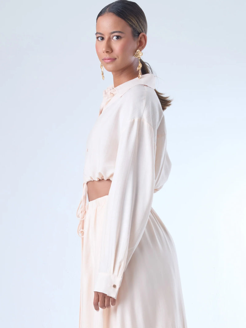 Blusa de corte relajado y largo cropped, con manga larga y puños con botones. Tiene cuello clásico camisero y una botonadura frontal. La prenda se ajusta a la cintura mediante un ligero fruncido con cordón interno, creando un efecto ablusado. Su estilo es casual con un toque sofisticado y femenino.

Detalles:

Color: Curuba

Material:18% Nylon 82% Rayón