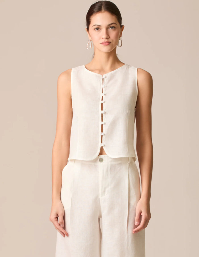 Blusa formal tipo chaleco en lino blanco perla, sin mangas y con escote redondo. Incluye cierre frontal con botones forrados y un dobladillo ligeramente curvado que realza la silueta. Una prenda fresca y elegante para looks profesionales o casual chic.