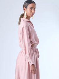 Blusa de corte relajado y largo cropped, con manga larga y puños con botones. Tiene cuello clásico camisero y una botonadura frontal. La prenda se ajusta a la cintura mediante un ligero fruncido con cordón interno, creando un efecto ablusado. Su estilo es casual con un toque sofisticado y femenino.

Detalles:

Color: Rosa