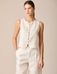 Top tipo chaleco en lino blanco perla, sin mangas y con escote redondo. Cierre frontal con botones forrados y diseño entallado en la parte superior. Su dobladillo curvado aporta un toque estructurado y sofisticado, ideal para outfits frescos y elegantes.