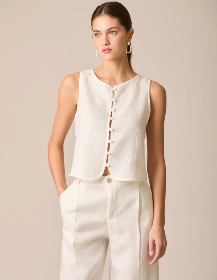 Top tipo chaleco en lino blanco perla, sin mangas y con escote redondo. Cierre frontal con botones forrados y diseño entallado en la parte superior. Su dobladillo curvado aporta un toque estructurado y sofisticado, ideal para outfits frescos y elegantes.