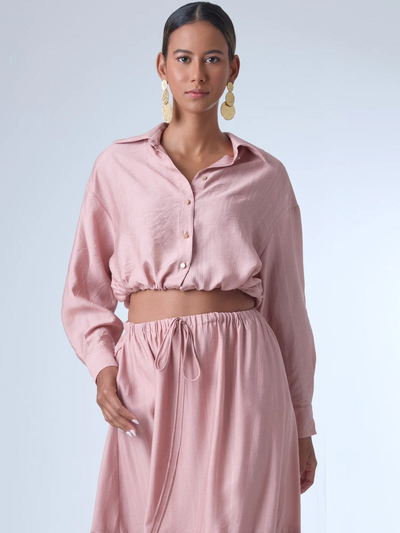 Blusa de corte relajado y largo cropped, con manga larga y puños con botones. Tiene cuello clásico camisero y una botonadura frontal. La prenda se ajusta a la cintura mediante un ligero fruncido con cordón interno, creando un efecto ablusado. Su estilo es casual con un toque sofisticado y femenino.

Detalles:

Color: Rosa