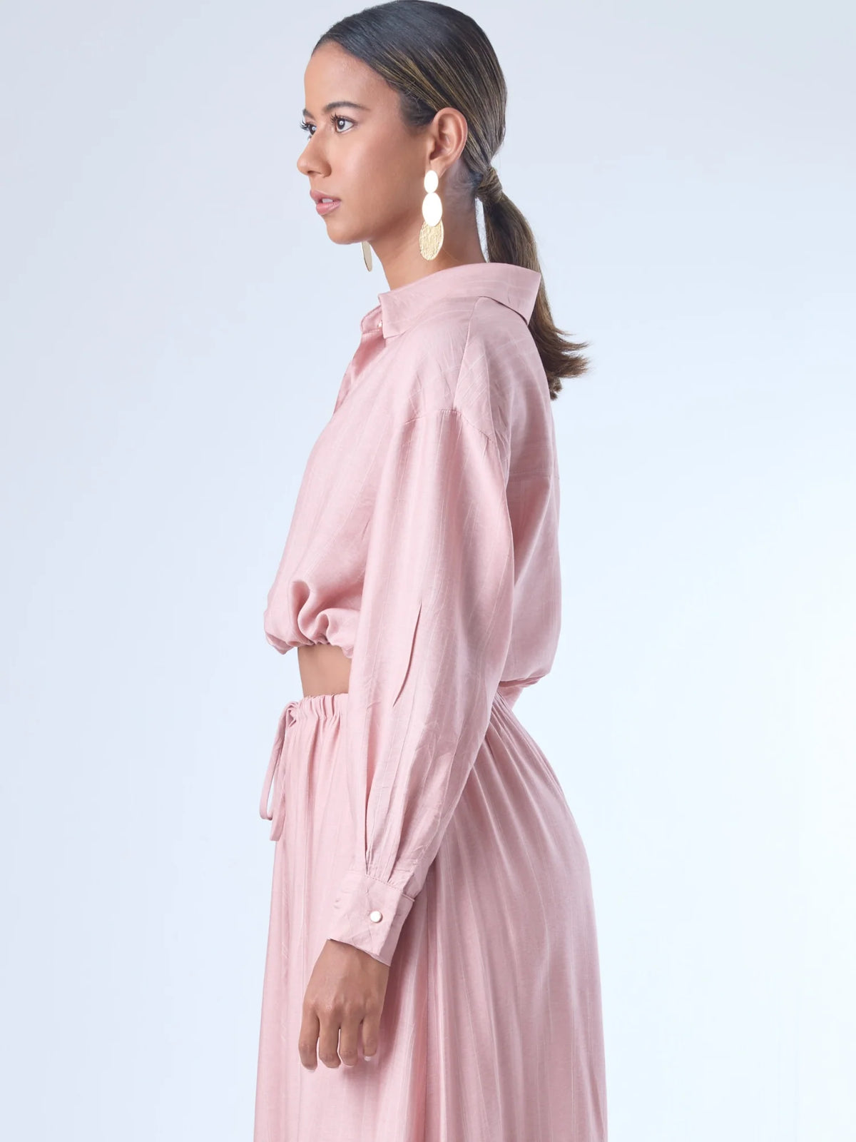 Blusa de corte relajado y largo cropped, con manga larga y puños con botones. Tiene cuello clásico camisero y una botonadura frontal. La prenda se ajusta a la cintura mediante un ligero fruncido con cordón interno, creando un efecto ablusado. Su estilo es casual con un toque sofisticado y femenino.

Detalles:

Color: Rosa