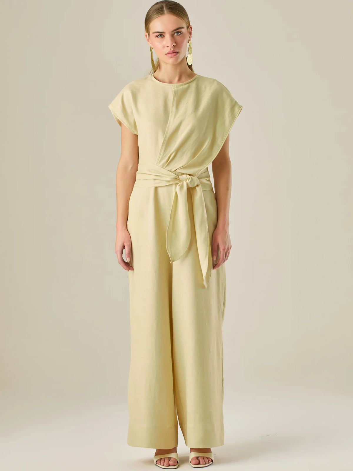 Este enterizo presenta un diseño elegante y minimalista, ideal para ocasiones casuales con estilo o eventos diurnos sofisticados. Confeccionado en un tono beige claro, tiene un corte relajado con mangas tipo kimono, cuello redondo y un diseño limpio sin botones ni cierres visibles en el frente. La prenda se ajusta en la cintura con un cinturón del mismo tejido, que se anuda al frente, creando una silueta favorecedora. El pantalón es de pierna ancha y caída fluida, lo que aporta comodidad y movimiento. Compl