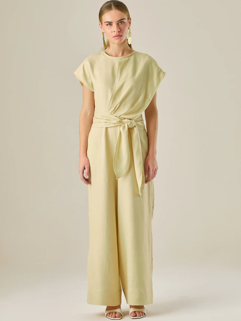 Este enterizo presenta un diseño elegante y minimalista, ideal para ocasiones casuales con estilo o eventos diurnos sofisticados. Confeccionado en un tono beige claro, tiene un corte relajado con mangas tipo kimono, cuello redondo y un diseño limpio sin botones ni cierres visibles en el frente. La prenda se ajusta en la cintura con un cinturón del mismo tejido, que se anuda al frente, creando una silueta favorecedora. El pantalón es de pierna ancha y caída fluida, lo que aporta comodidad y movimiento. Compl