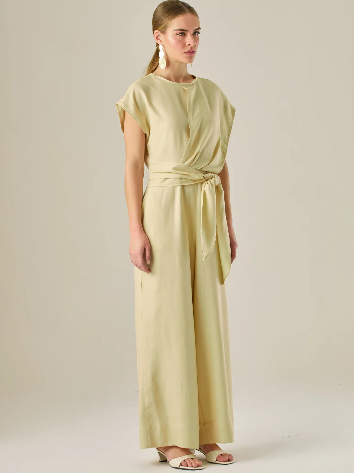 Este enterizo presenta un diseño elegante y minimalista, ideal para ocasiones casuales con estilo o eventos diurnos sofisticados. Confeccionado en un tono beige claro, tiene un corte relajado con mangas tipo kimono, cuello redondo y un diseño limpio sin botones ni cierres visibles en el frente. La prenda se ajusta en la cintura con un cinturón del mismo tejido, que se anuda al frente, creando una silueta favorecedora. El pantalón es de pierna ancha y caída fluida, lo que aporta comodidad y movimiento. Compl