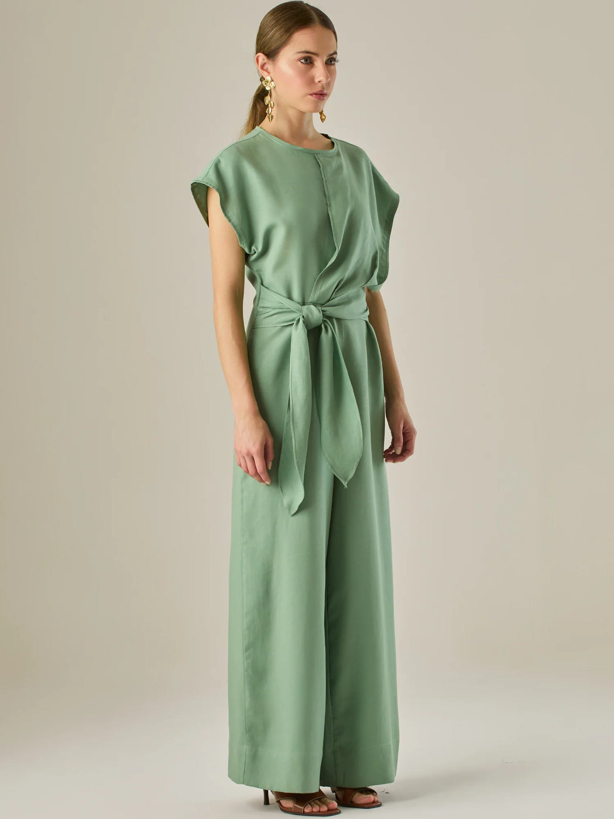 Este enterizo presenta un diseño elegante y minimalista, ideal para ocasiones casuales con estilo o eventos diurnos sofisticados. Confeccionado en un tono beige claro, tiene un corte relajado con mangas tipo kimono, cuello redondo y un diseño limpio sin botones ni cierres visibles en el frente. La prenda se ajusta en la cintura con un cinturón del mismo tejido, que se anuda al frente, creando una silueta favorecedora. El pantalón es de pierna ancha y caída fluida, lo que aporta comodidad y movimiento. Compl