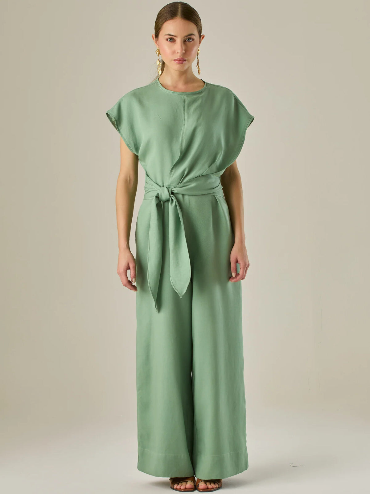 Este enterizo presenta un diseño elegante y minimalista, ideal para ocasiones casuales con estilo o eventos diurnos sofisticados. Confeccionado en un tono beige claro, tiene un corte relajado con mangas tipo kimono, cuello redondo y un diseño limpio sin botones ni cierres visibles en el frente. La prenda se ajusta en la cintura con un cinturón del mismo tejido, que se anuda al frente, creando una silueta favorecedora. El pantalón es de pierna ancha y caída fluida, lo que aporta comodidad y movimiento. Compl