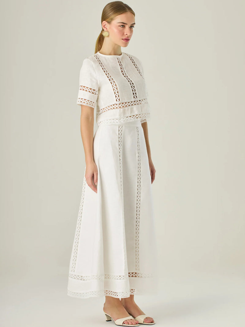 Esta elegante falda larga está confeccionada en un tejido blanco de apariencia ligera y fresca, ideal para climas cálidos o estilos veraniegos. Su diseño de silueta A le da una caída fluida y femenina, que aporta comodidad sin perder sofisticación. La falda destaca por sus aplicaciones de encaje calado en líneas verticales y una franja decorativa en el bajo, que añade un aire romántico y artesanal a la prenda. La cintura alta estiliza la figura y permite combinarla fácilmente con tops por dentro. Es perfect