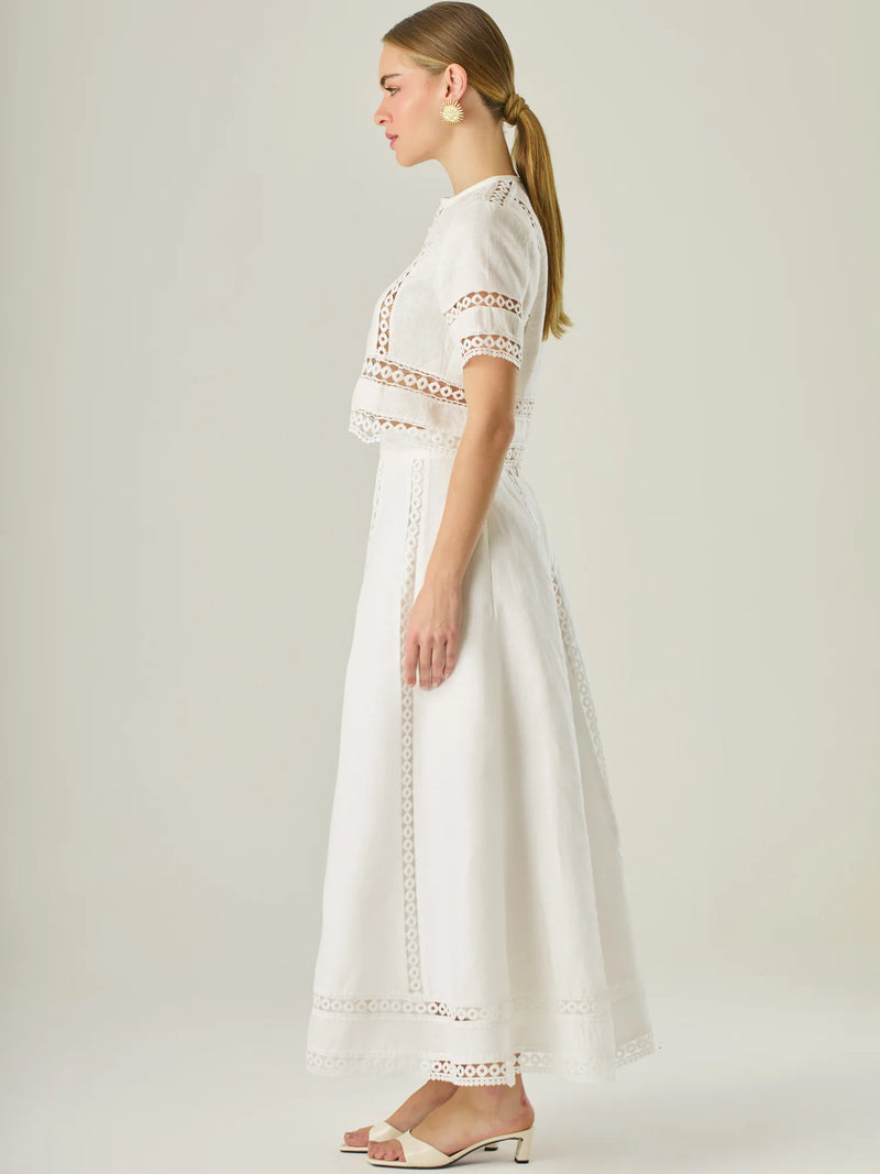 Esta elegante falda larga está confeccionada en un tejido blanco de apariencia ligera y fresca, ideal para climas cálidos o estilos veraniegos. Su diseño de silueta A le da una caída fluida y femenina, que aporta comodidad sin perder sofisticación. La falda destaca por sus aplicaciones de encaje calado en líneas verticales y una franja decorativa en el bajo, que añade un aire romántico y artesanal a la prenda. La cintura alta estiliza la figura y permite combinarla fácilmente con tops por dentro. Es perfect