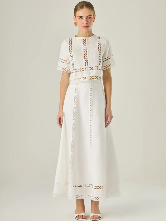 Esta elegante falda larga está confeccionada en un tejido blanco de apariencia ligera y fresca, ideal para climas cálidos o estilos veraniegos. Su diseño de silueta A le da una caída fluida y femenina, que aporta comodidad sin perder sofisticación. La falda destaca por sus aplicaciones de encaje calado en líneas verticales y una franja decorativa en el bajo, que añade un aire romántico y artesanal a la prenda. La cintura alta estiliza la figura y permite combinarla fácilmente con tops por dentro. Es perfect