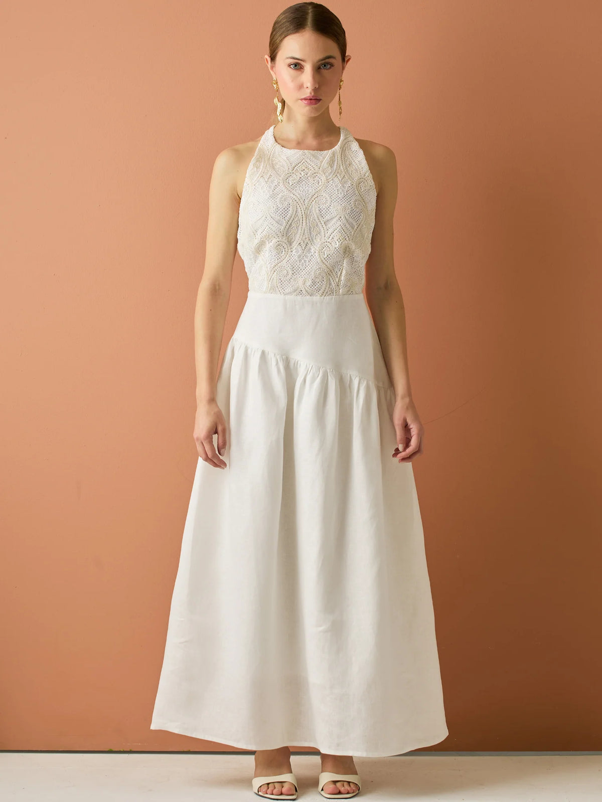 Vestido largo sin mangas con escote halter y cuerpo bordado en delicado encaje. La falda de lino con caída fluida aporta frescura y movimiento, creando una silueta femenina y elegante ideal para ocasiones especiales con un aire natural y sofisticado.