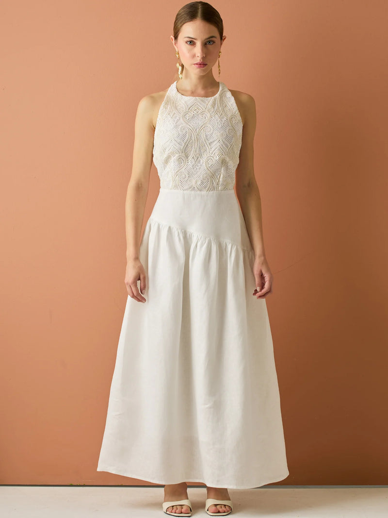Vestido largo sin mangas con escote halter y cuerpo bordado en delicado encaje. La falda de lino con caída fluida aporta frescura y movimiento, creando una silueta femenina y elegante ideal para ocasiones especiales con un aire natural y sofisticado.