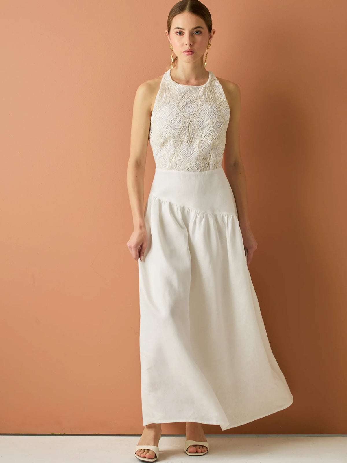 Vestido largo sin mangas con escote halter y cuerpo bordado en delicado encaje. La falda de lino con caída fluida aporta frescura y movimiento, creando una silueta femenina y elegante ideal para ocasiones especiales con un aire natural y sofisticado.