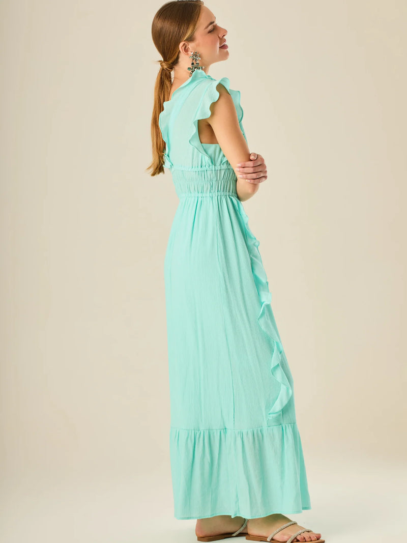 Vestido largo sin mangas con escote en "V" profundo y un diseño con volantes que recorren tanto los tirantes como la parte frontal del busto y la falda. La cintura está fruncida con elástico para dar definición y comodidad, mientras que la falda fluye suavemente con un detalle de volante en el dobladillo, aportando movimiento y un aire femenino al conjunto.