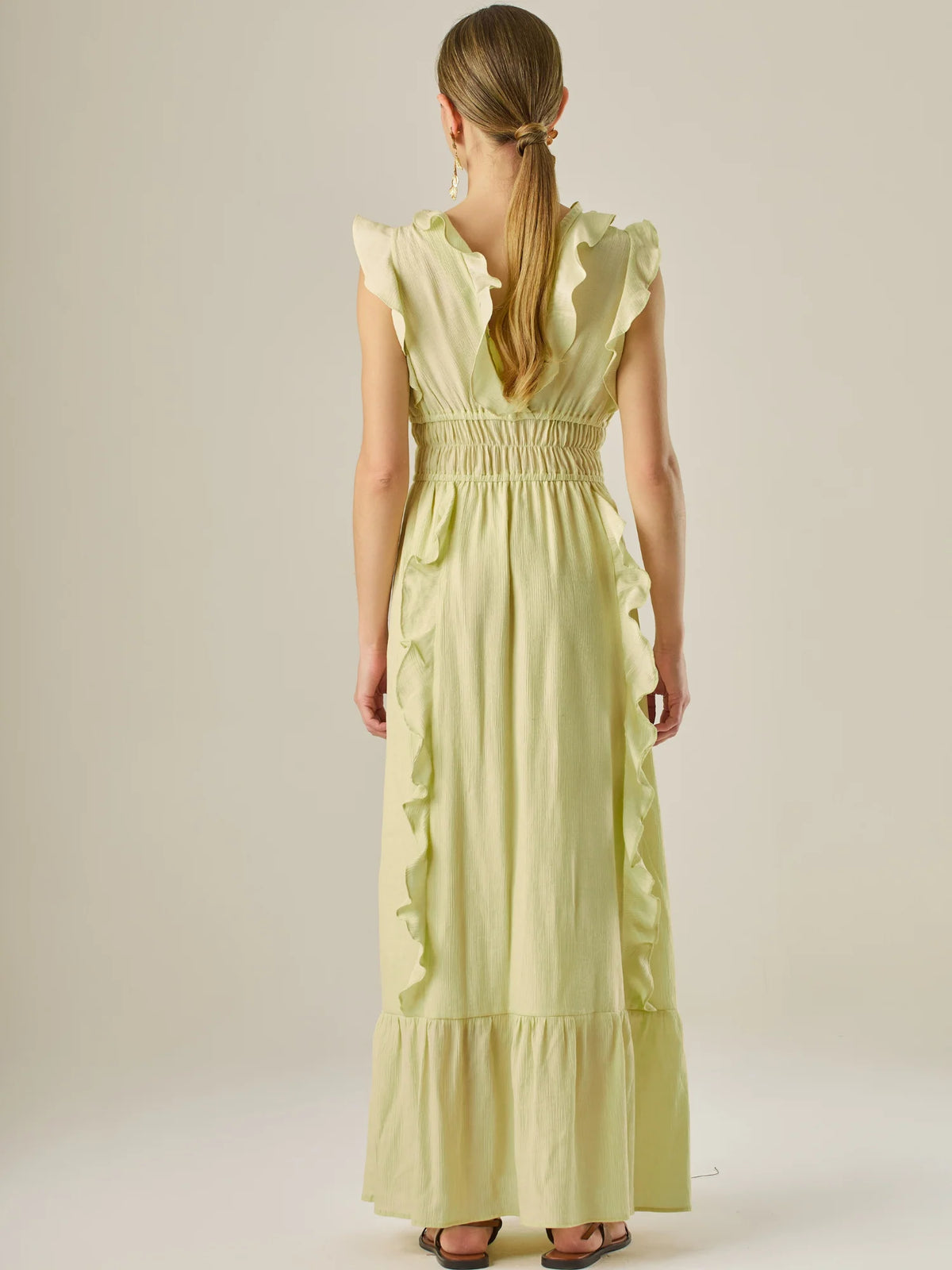 Vestido largo sin mangas con escote en "V" profundo y un diseño con volantes que recorren tanto los tirantes como la parte frontal del busto y la falda. La cintura está fruncida con elástico para dar definición y comodidad, mientras que la falda fluye suavemente con un detalle de volante en el dobladillo, aportando movimiento y un aire femenino al conjunto.