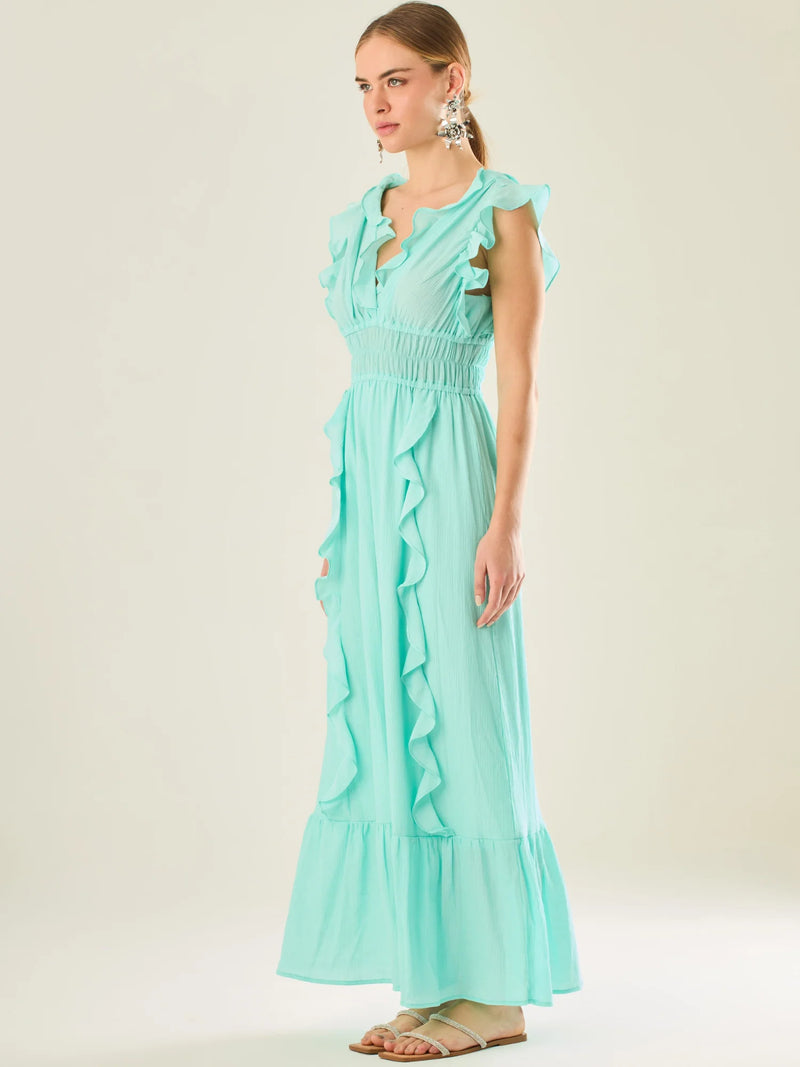 Vestido largo sin mangas con escote en "V" profundo y un diseño con volantes que recorren tanto los tirantes como la parte frontal del busto y la falda. La cintura está fruncida con elástico para dar definición y comodidad, mientras que la falda fluye suavemente con un detalle de volante en el dobladillo, aportando movimiento y un aire femenino al conjunto.