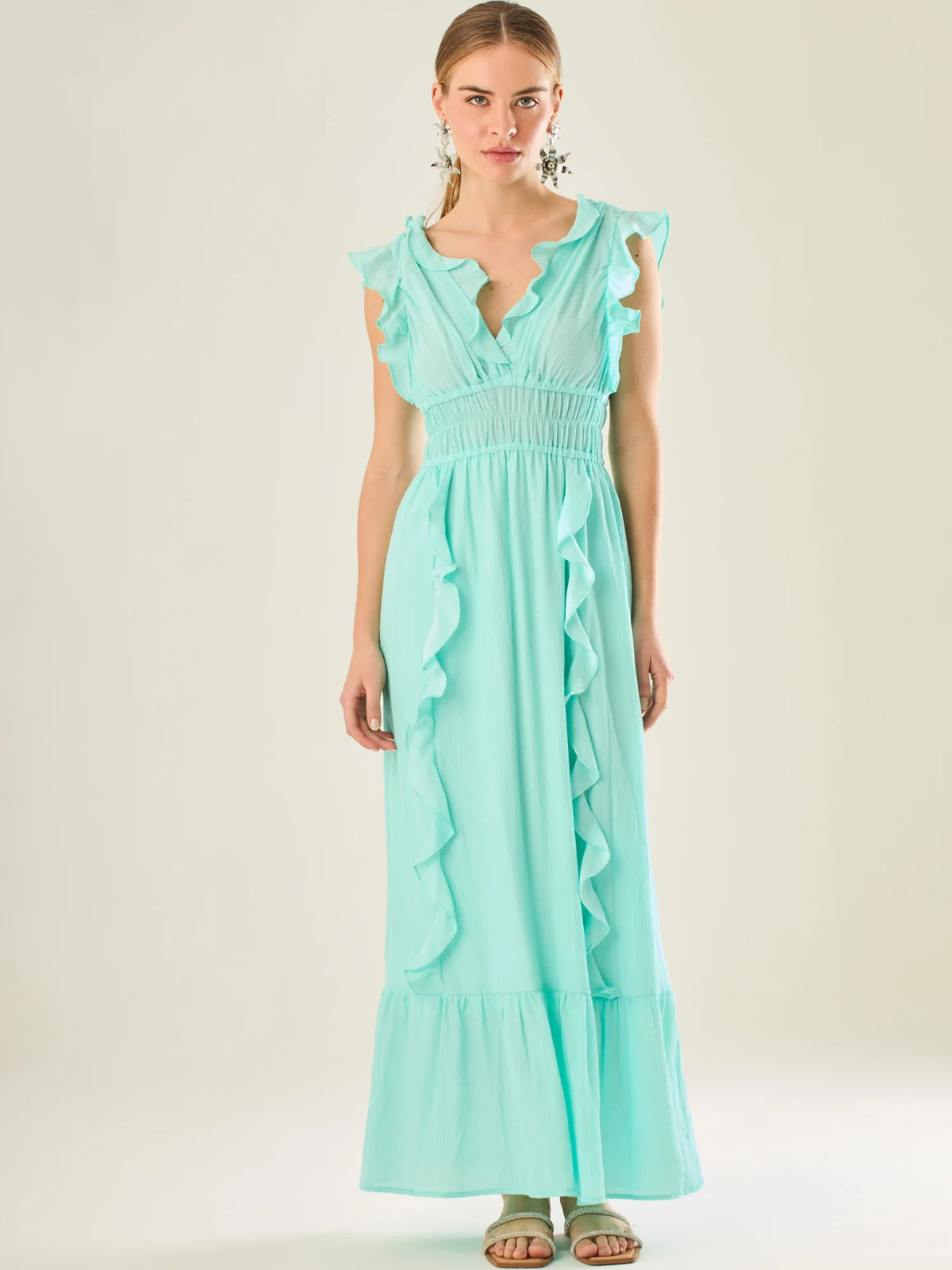 Vestido largo sin mangas con escote en "V" profundo y un diseño con volantes que recorren tanto los tirantes como la parte frontal del busto y la falda. La cintura está fruncida con elástico para dar definición y comodidad, mientras que la falda fluye suavemente con un detalle de volante en el dobladillo, aportando movimiento y un aire femenino al conjunto.