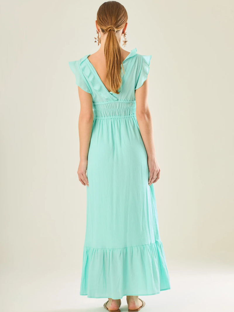 Vestido largo sin mangas con escote en "V" profundo y un diseño con volantes que recorren tanto los tirantes como la parte frontal del busto y la falda. La cintura está fruncida con elástico para dar definición y comodidad, mientras que la falda fluye suavemente con un detalle de volante en el dobladillo, aportando movimiento y un aire femenino al conjunto.